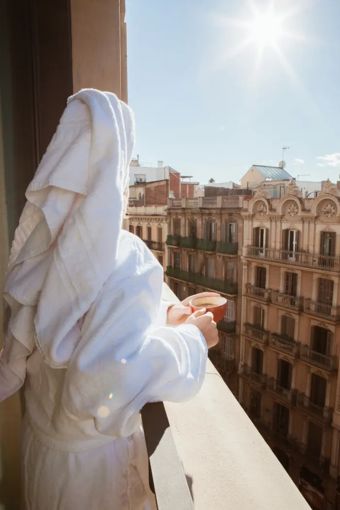 Una persona con bata blanca se sienta en un balcón con luz de sol y edificios al fondo.