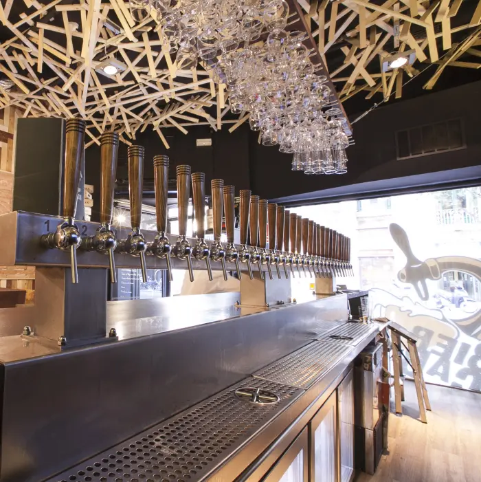 Se ve una barra de bar con grifos metálicos, botellas y una lámpara colgante decorativa en el techo.