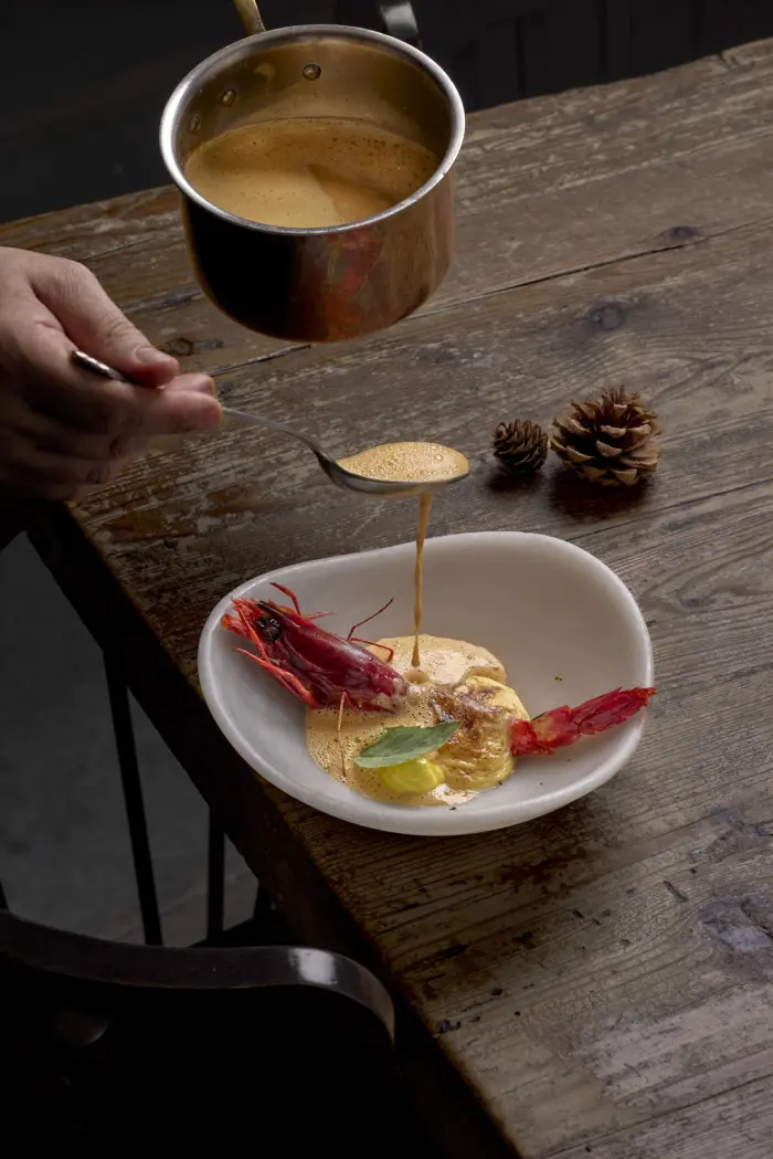 Una mano sostiene una taza de café junto a un plato con langostinos sobre una mesa de madera.
