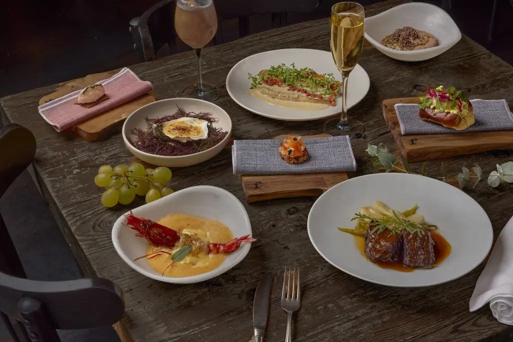 Se ve una mesa con varios platos servidos, pan, uvas y vasos con bebidas sobre manteles individuales.