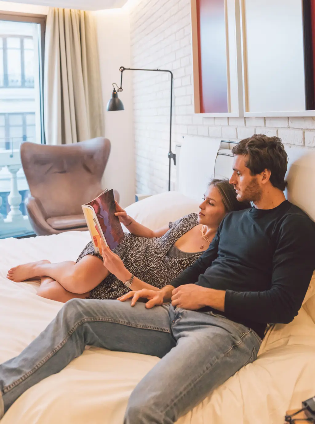 Una pareja se recuesta en una cama dentro de una habitación luminosa con ventana y cortinas beige.