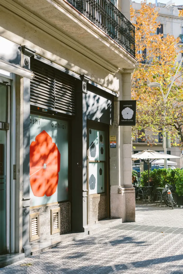 Se ve una fachada de edificio con una puerta de cristal, cartelera y una calle peatonal con mesas al aire libre.
