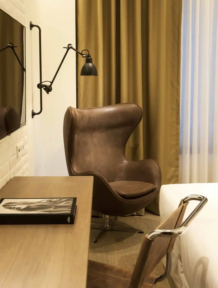 Se ve un escritorio con silla de oficina, una lámpara de lectura y cortinas junto a una cama en un dormitorio.