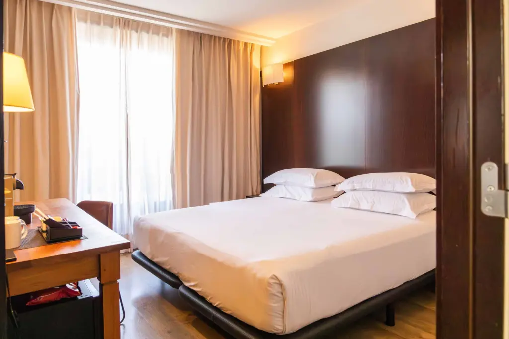 Se ve una cama doble con cojines blancos junto a una mesita de noche y cortinas claras.