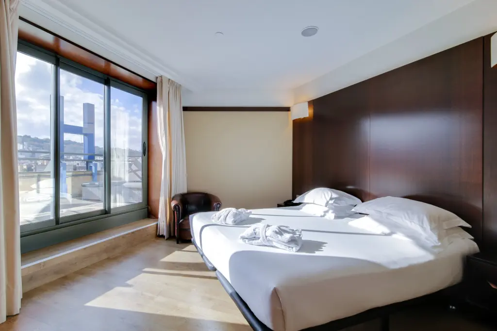 Se ve una cama doble, mesita y un ventanal con cortinas en una habitación luminosa.