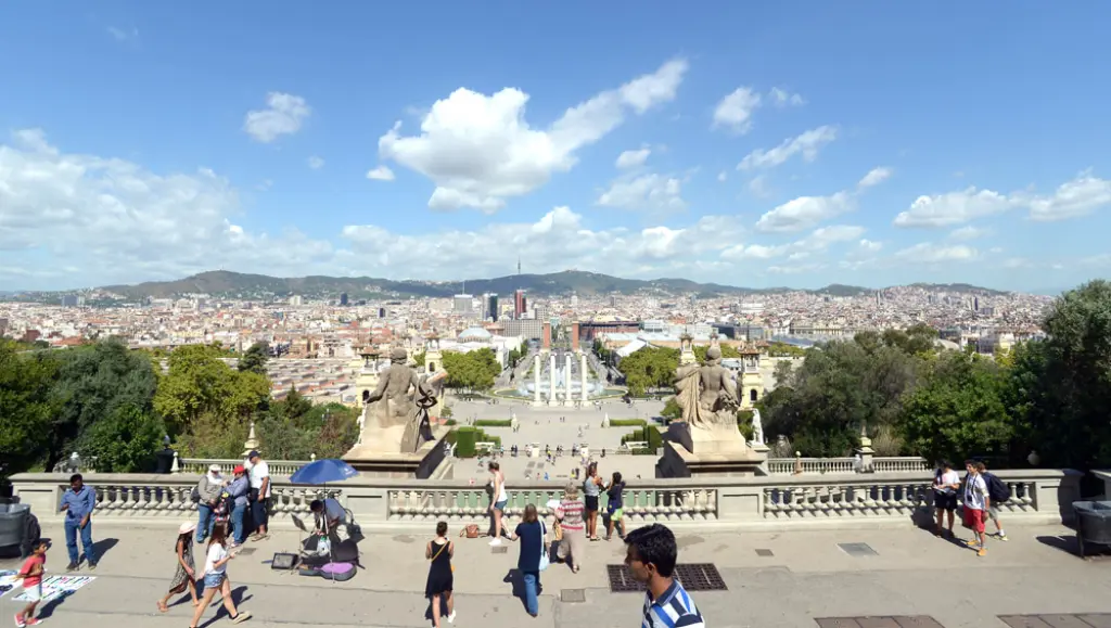 Personas pasean en un mirador de piedra con escaleras y la ciudad al fondo bajo un cielo despejado.